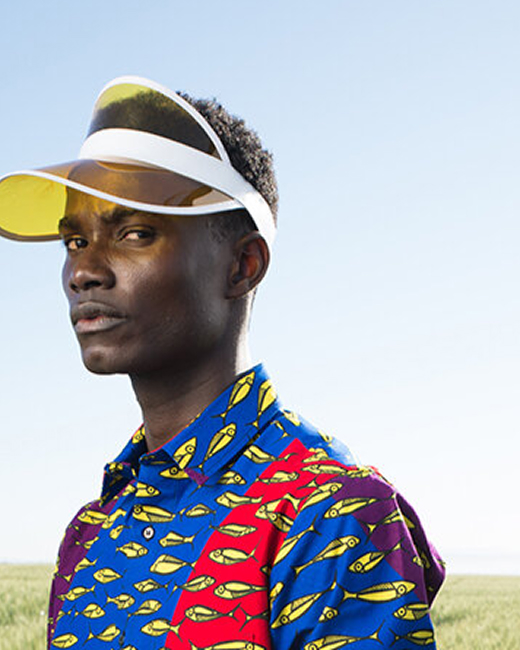 A young Black man wearing a sun visor and a blue and red shirt is facing slighty left of the camera looking at the viewer