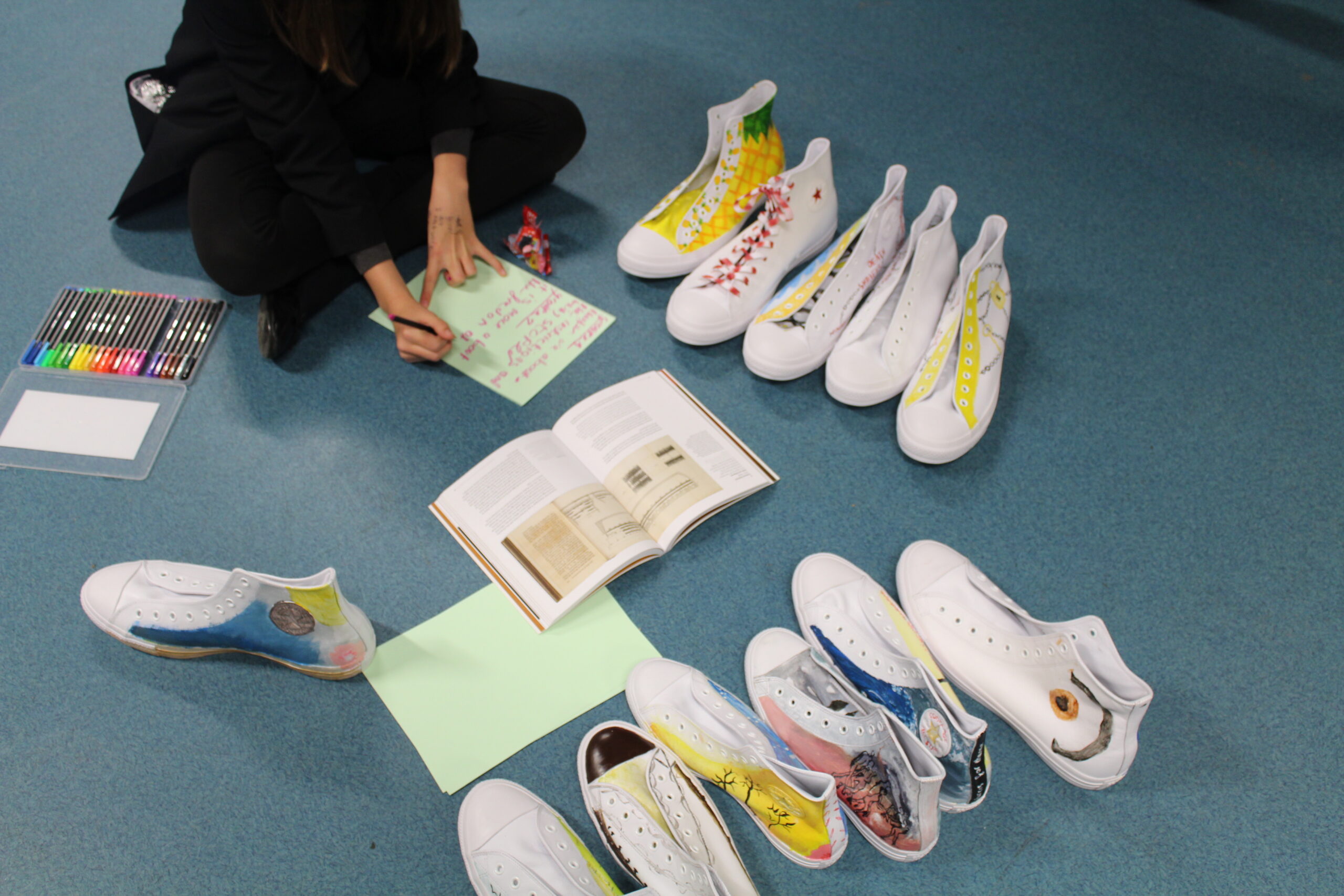 A young person sitting on the floor next to pairs of hand-coloured trainers