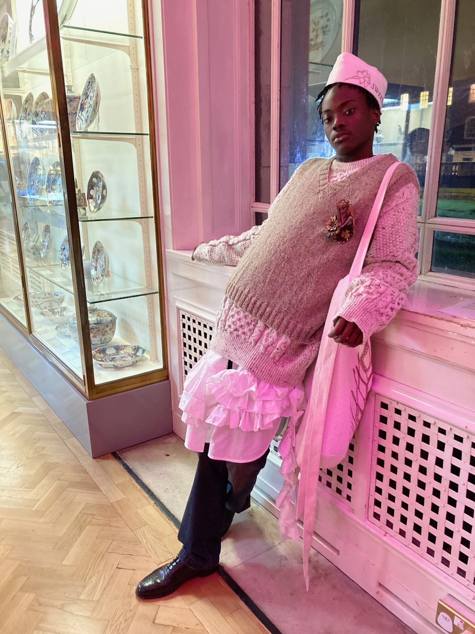 A young Black man in high fashion leaning against a wall in a museum